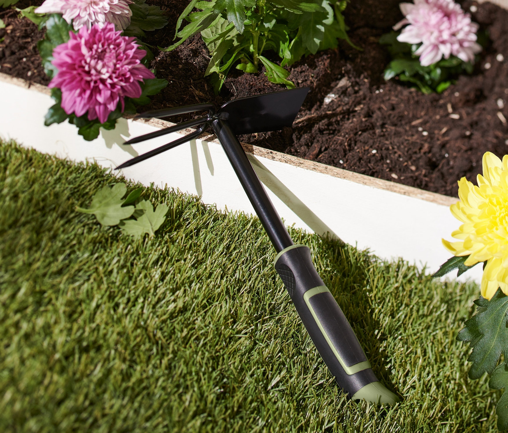 Siyah çift taraflı bahçe çapası, sarı çiçekli bir saksının yanında çimenlerin üzerinde duruyor.