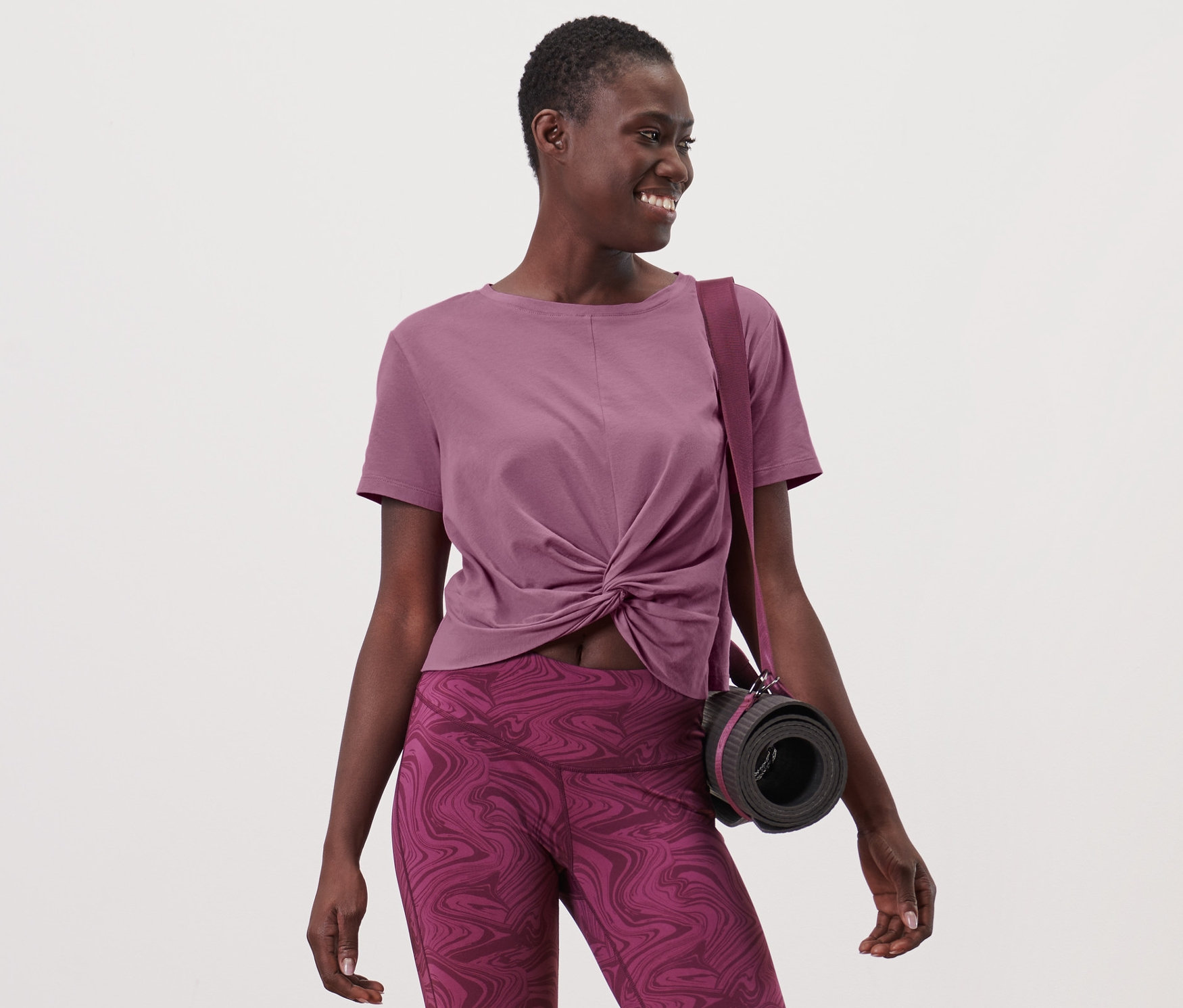 Kadın spor tişörtü ve tayt giyiyor, omzunda çantası ve yoga matı var.