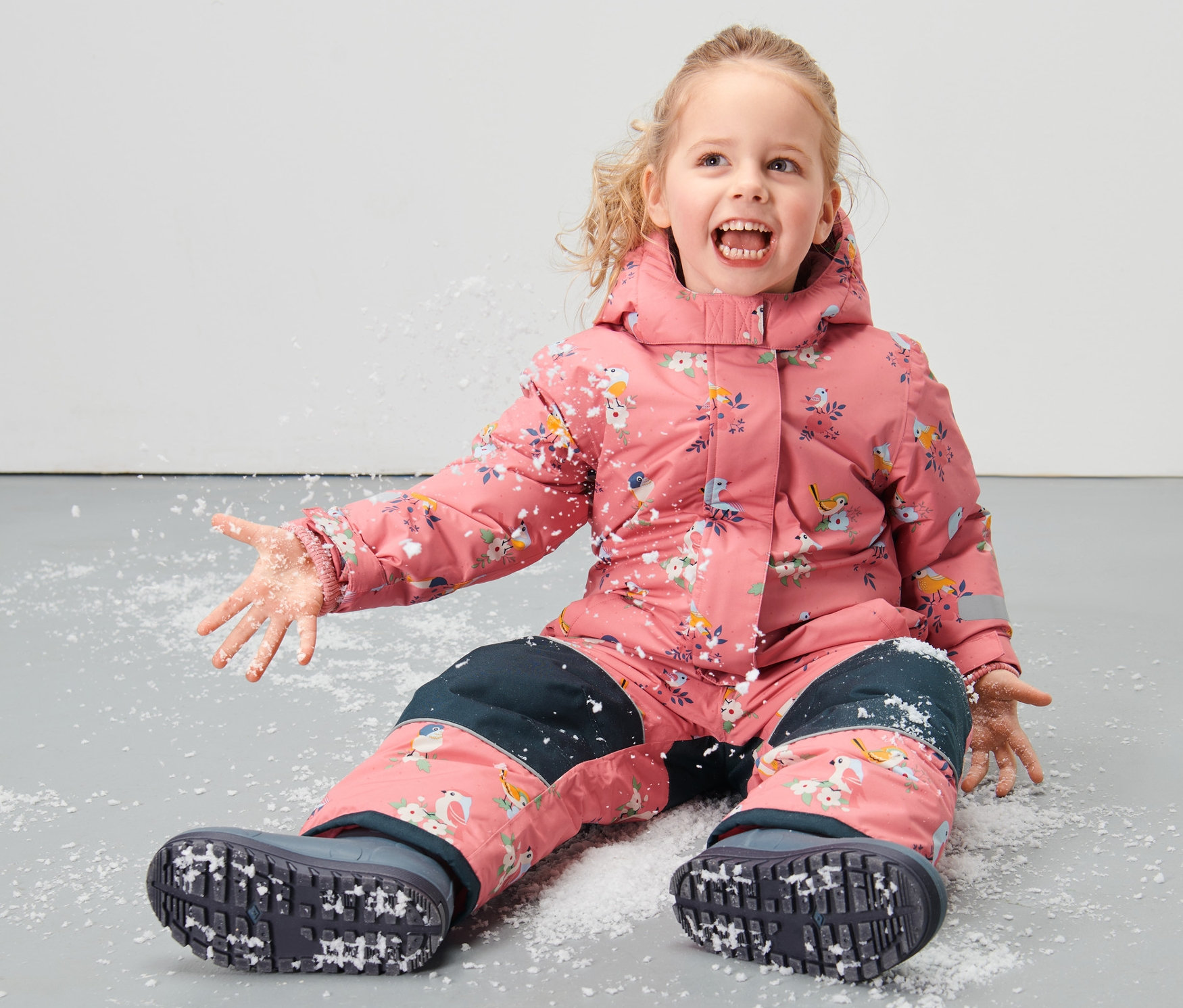 Gülümseyen sarışın bir çocuk yerde oturmuş, üzerinde kuş baskıları olan küçük çocuk kar tulumuyla karda oynuyor.