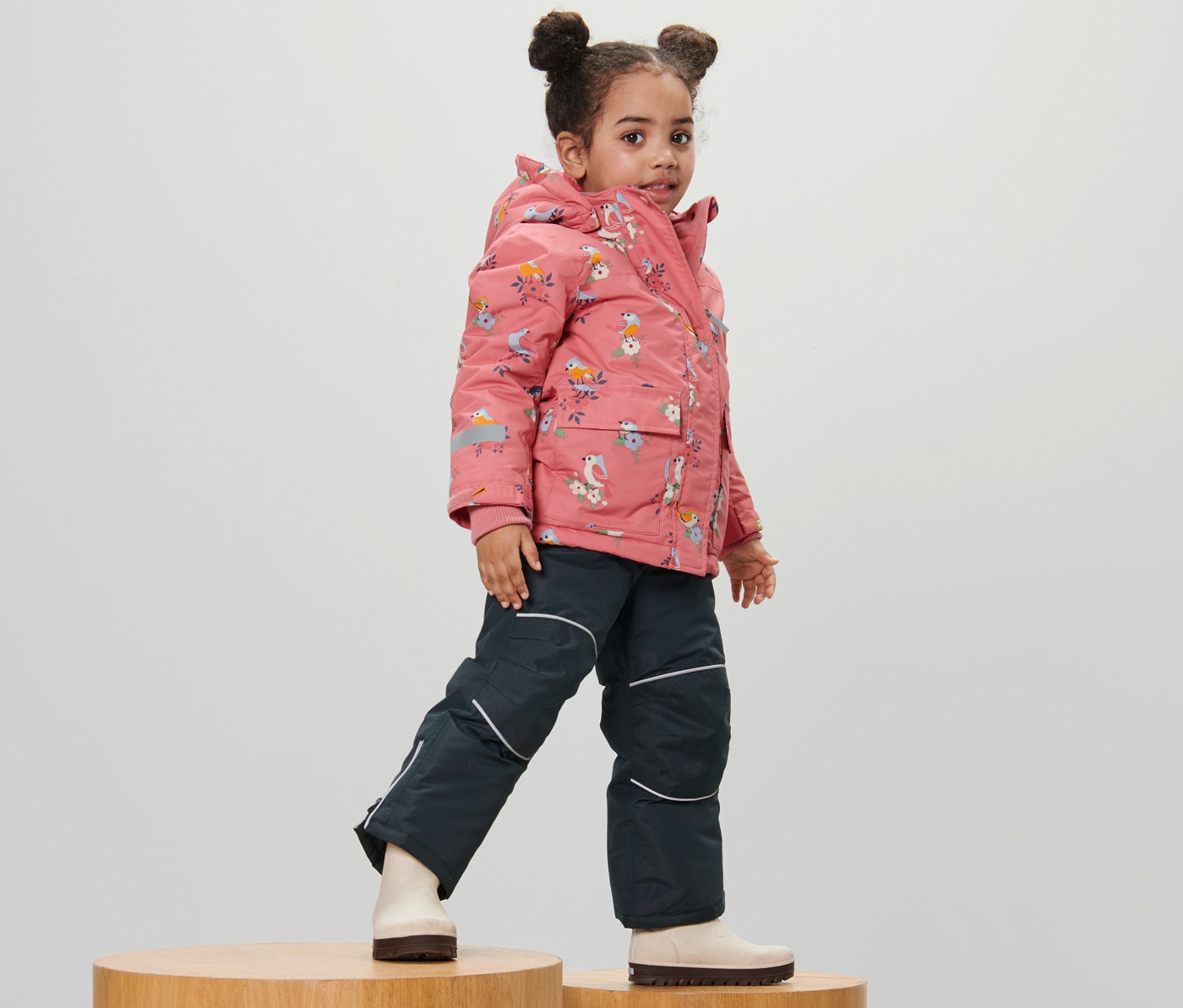 Kız çocuk, tamamı kuş baskılı 2 parçalı kar kıyafetiyle ahşap yükseltiler üzerinde poz veriyor.