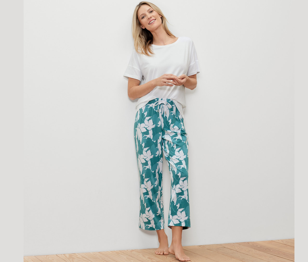 Sarışın bir kadın beyaz bir duvara yaslanmış, beyaz bir tişört ve desenli yeşil bir pijama altı giyiyor.