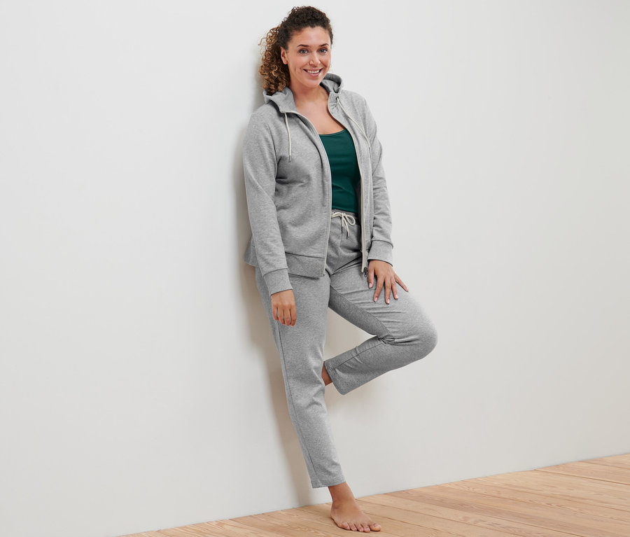 Duvara yaslanmış bir kadın gri Sweat Ceket, yeşil ve kırık beyaz 2 Adet Atlet ve gri Sweat Pantolon giyiyor.