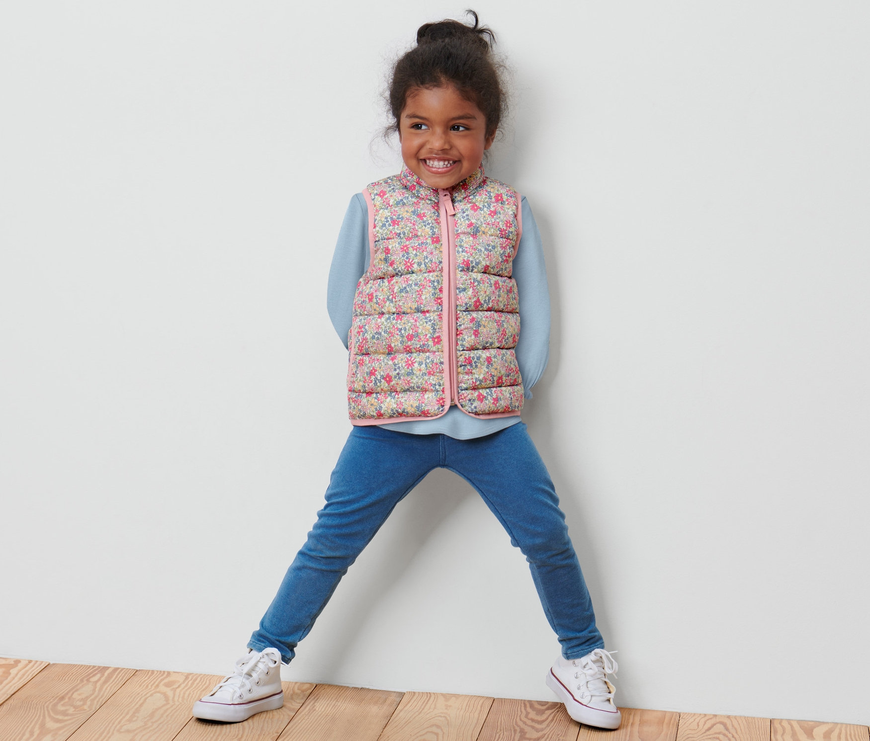 Desenli kapitone yelek Küçük Çocuk Kapitone Yelek, Çiçekli, mavi kazak, kot pantolon ve spor ayakkabı giyen bir kız çocuğu poz veriyor.