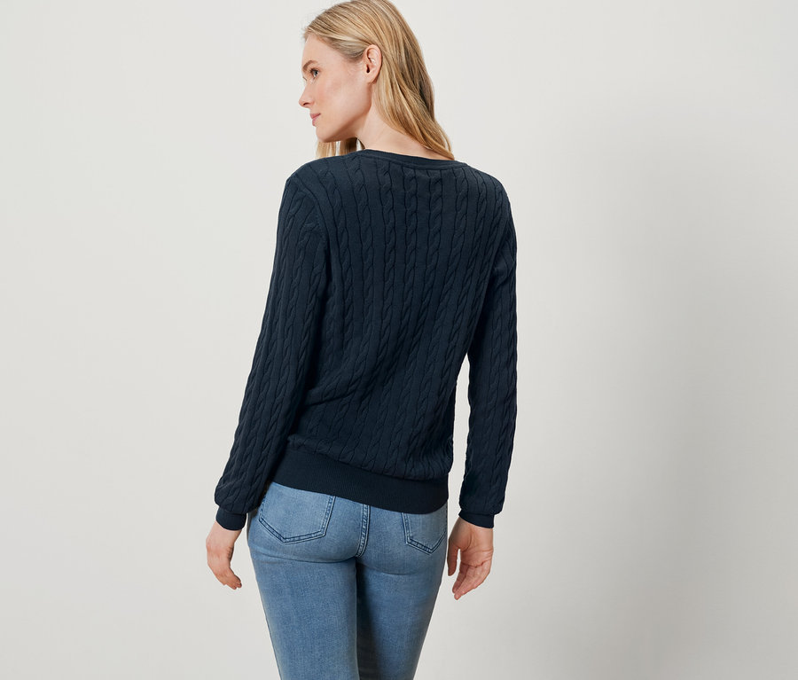 Sarışın saçlı, mavi bir kazak ve kot pantolon giymiş bir kadın kameraya arkası dönük duruyor.