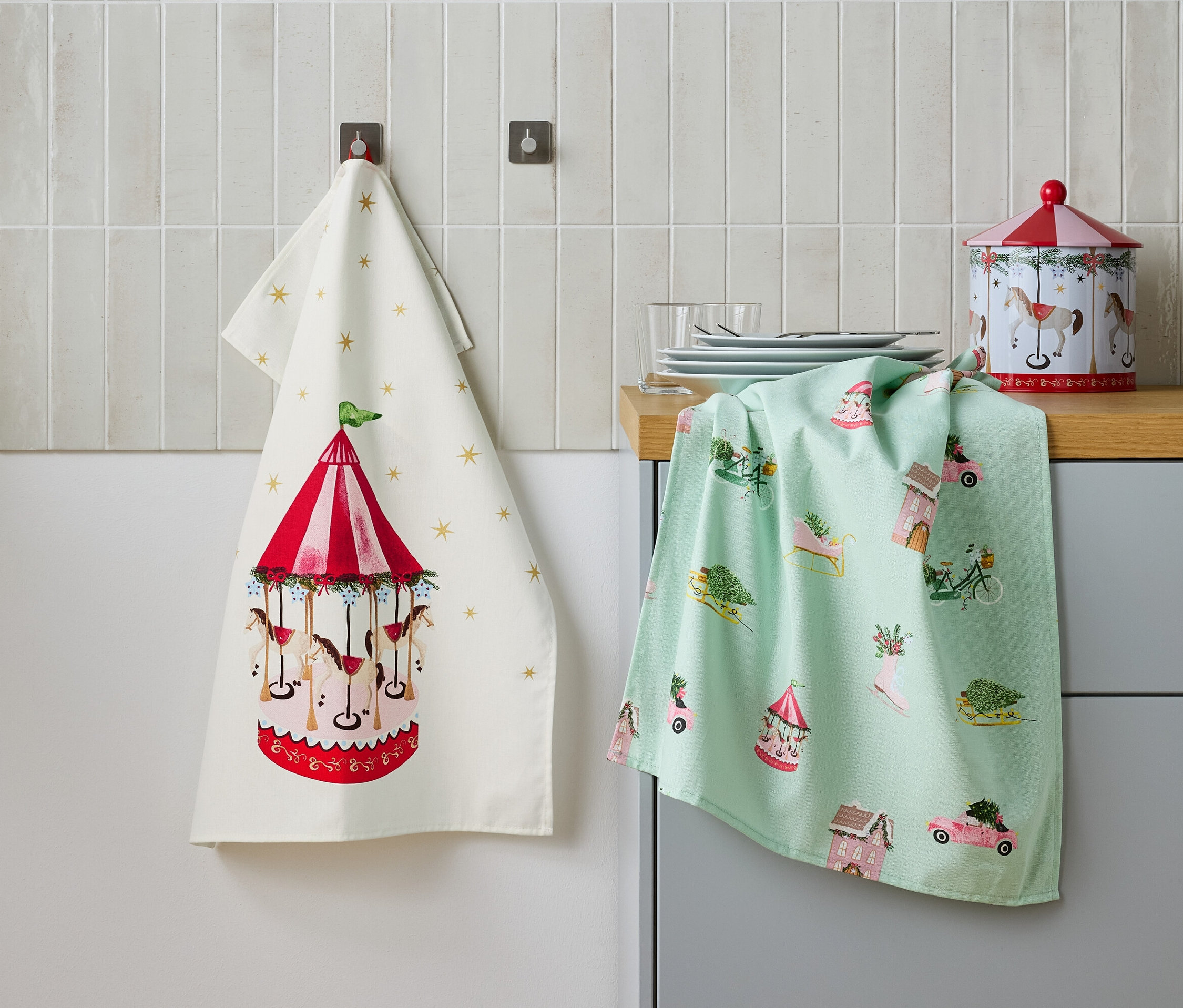 İki mutfak havlusu yan yana duvarda asılı duruyor, biri atlı karınca deseniyle, diğeri ise şenlikli motiflerle, ayrıca masanın üzerinde metal bir kutu bulunuyor.