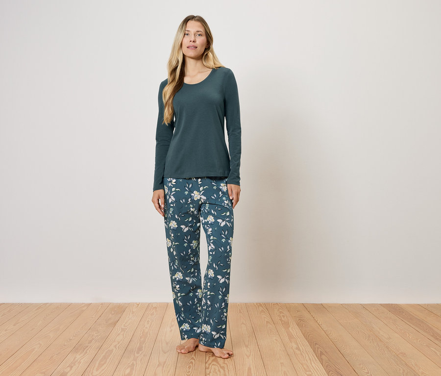 Bir kadın ayakta duruyor, üzerinde uzun kollu, koyu yeşil bir pijama ve çiçekli bir pantolon var.