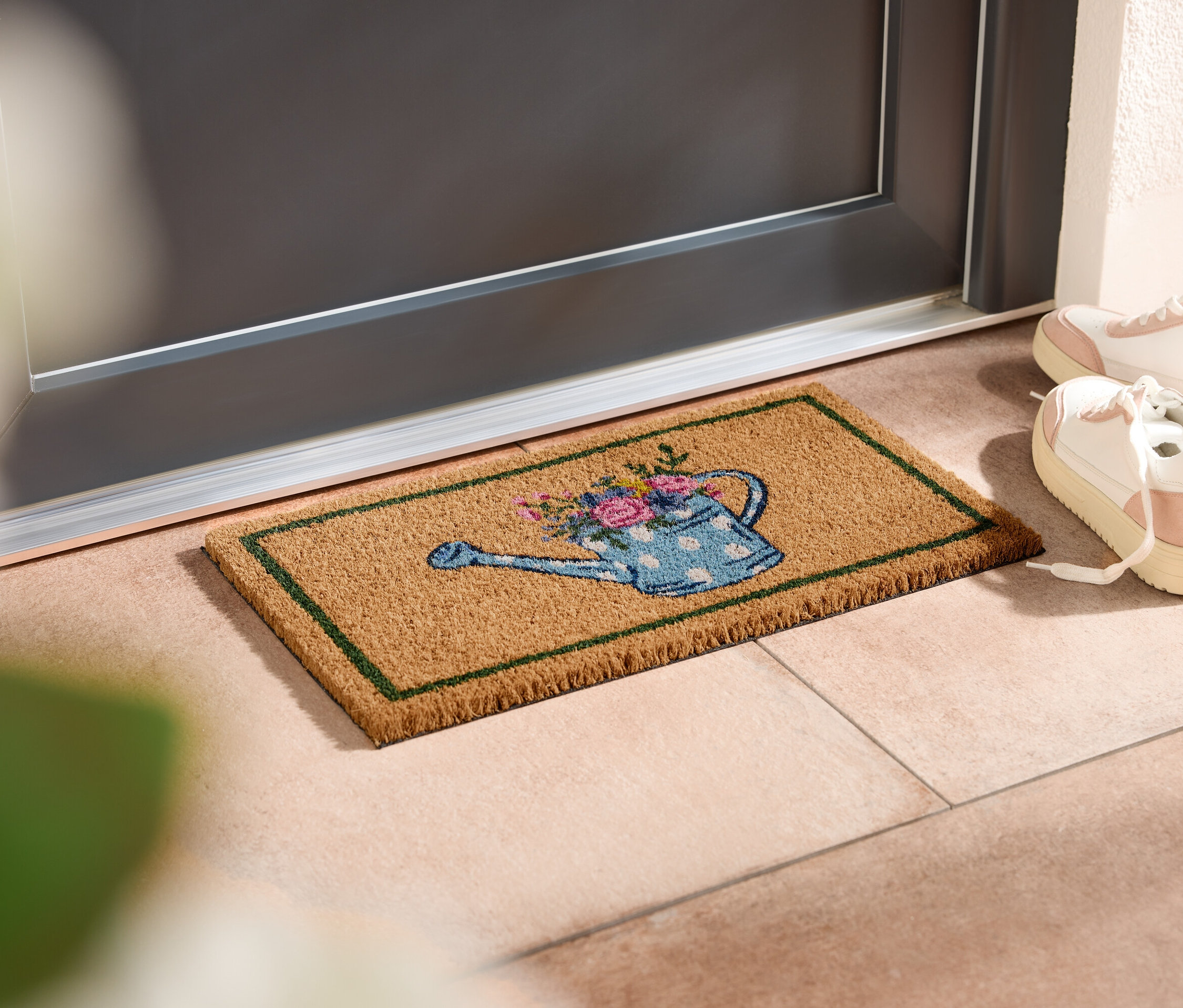 Kapı önünde sulama kabı desenli paspas, yanında beyaz spor ayakkabılar duruyor.