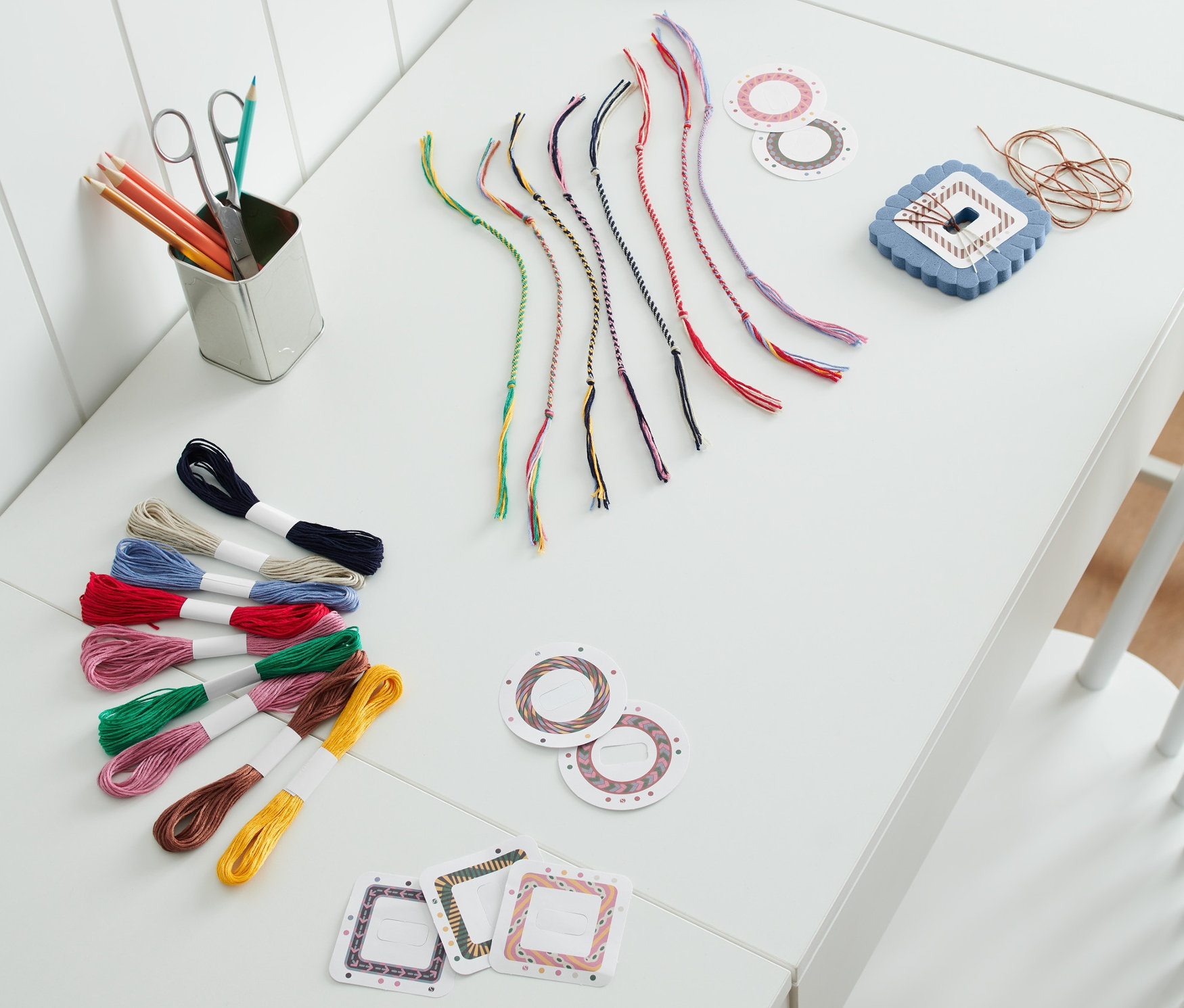 Beyaz bir masanın üzerinde El İşi Seti »Arkadaşlık Bilekliği«, renkli ipler ve mavi bir lamba bulunmaktadır.