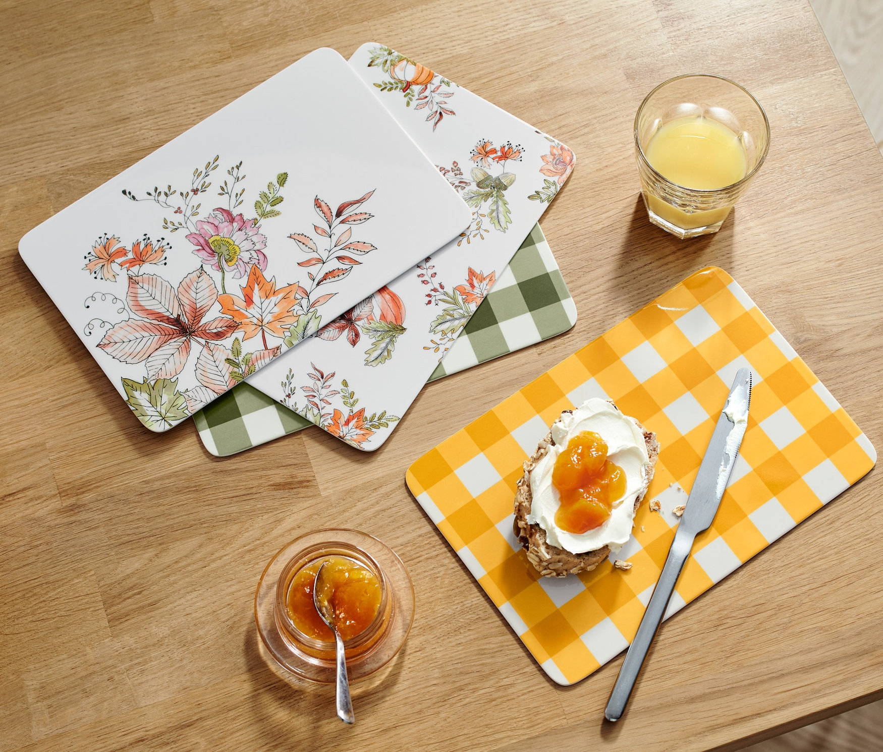 Dört adet kahvaltı servis tahtası, bir bardak meyve suyunun, reçelli ekmeğin, reçelin ve bir sepet ekmeğin yanında ahşap bir masa üzerinde duruyor.