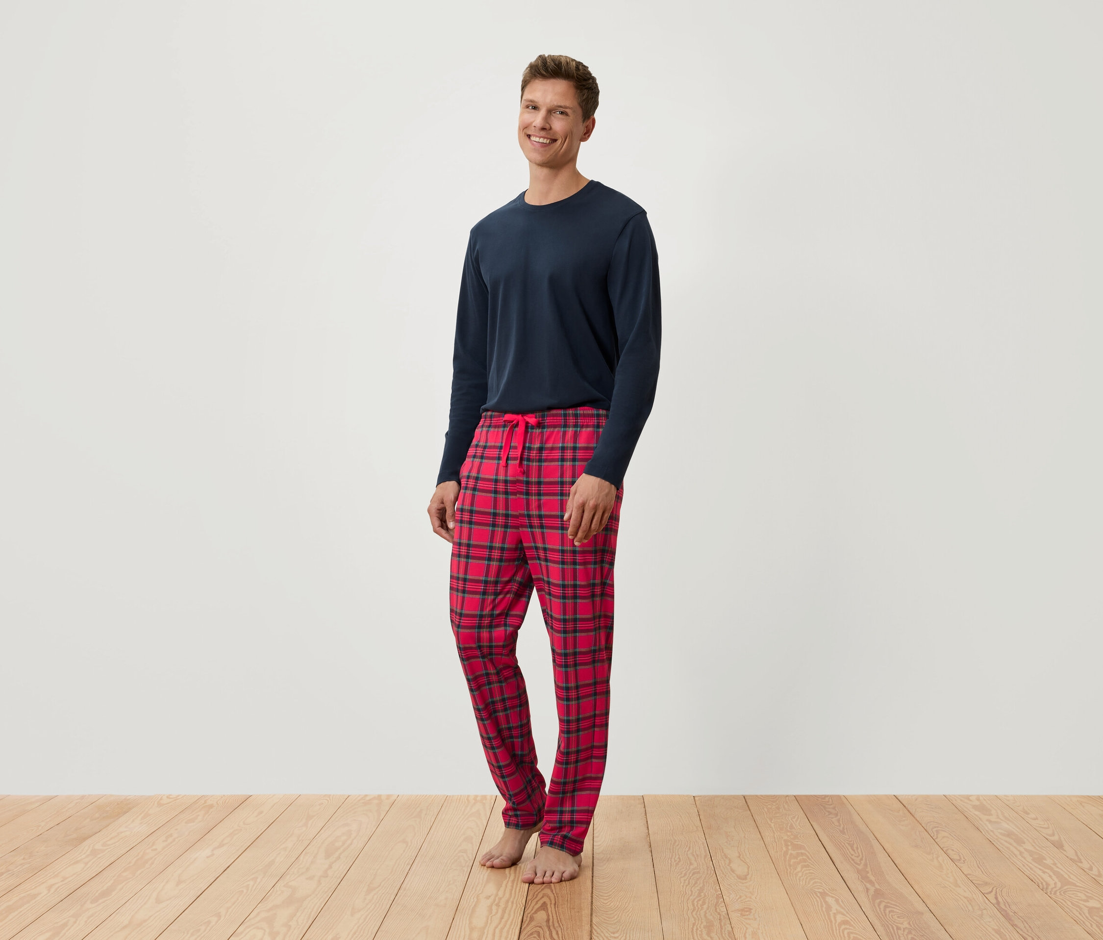 Adam ayakta duruyor, uzun kollu lacivert bir tişört ve kırmızı kareli bir pijama altı giyiyor.