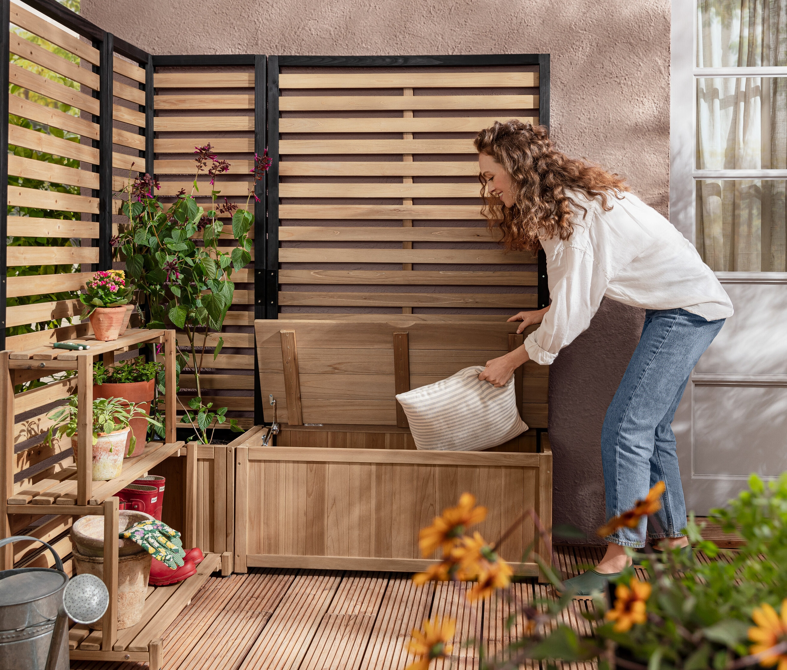 Bir kadın Sandıklı Banklı Paravan Modülü »Elin« içine bir yastık koyuyor. Arka planda Saksılı Paravan Modülü »Elin« ve Raflı Paravan Modülü »Elin« bulunuyor.