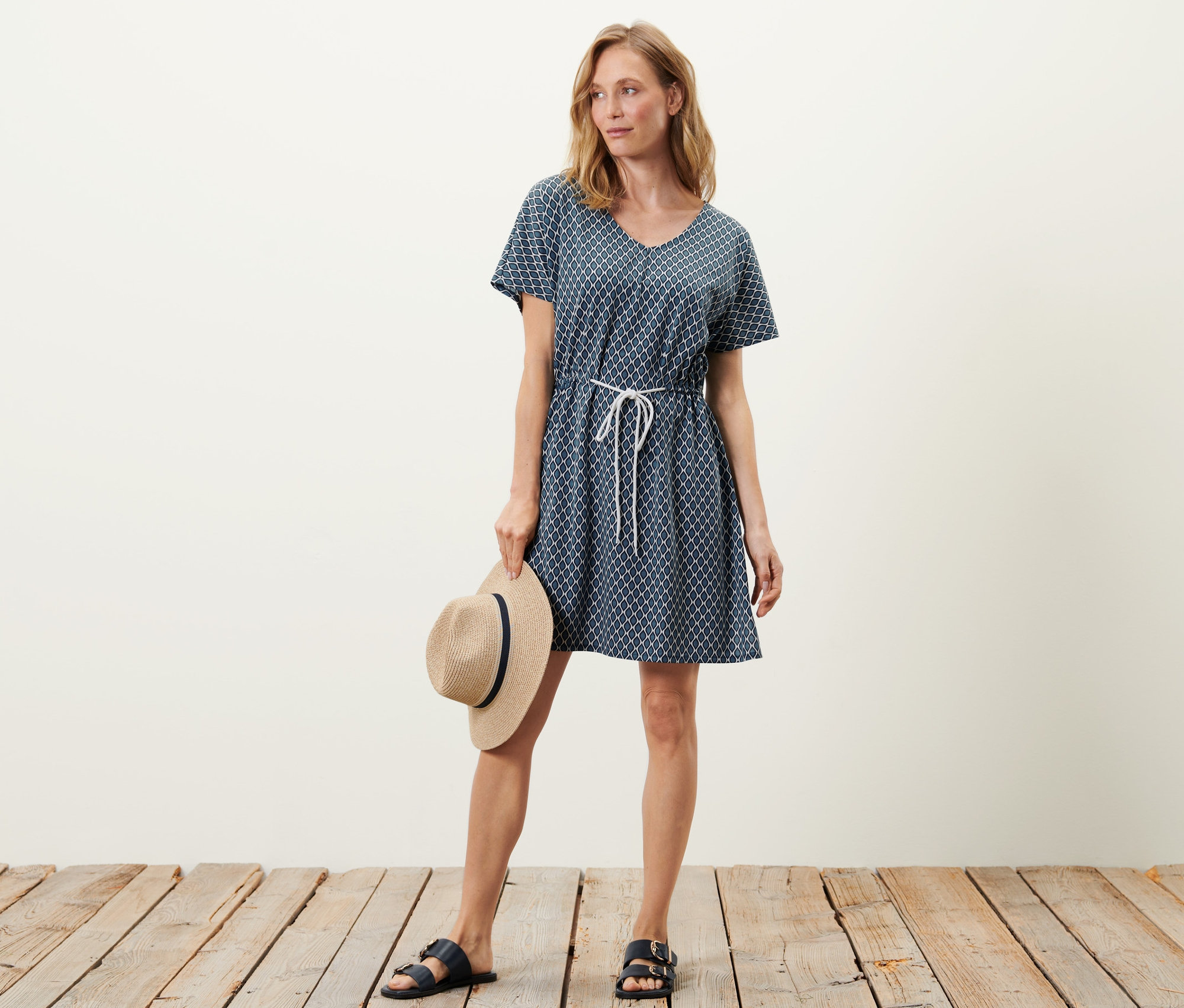 Kadın plaj elbisesiyle poz veriyor ve bir şapka tutuyor.