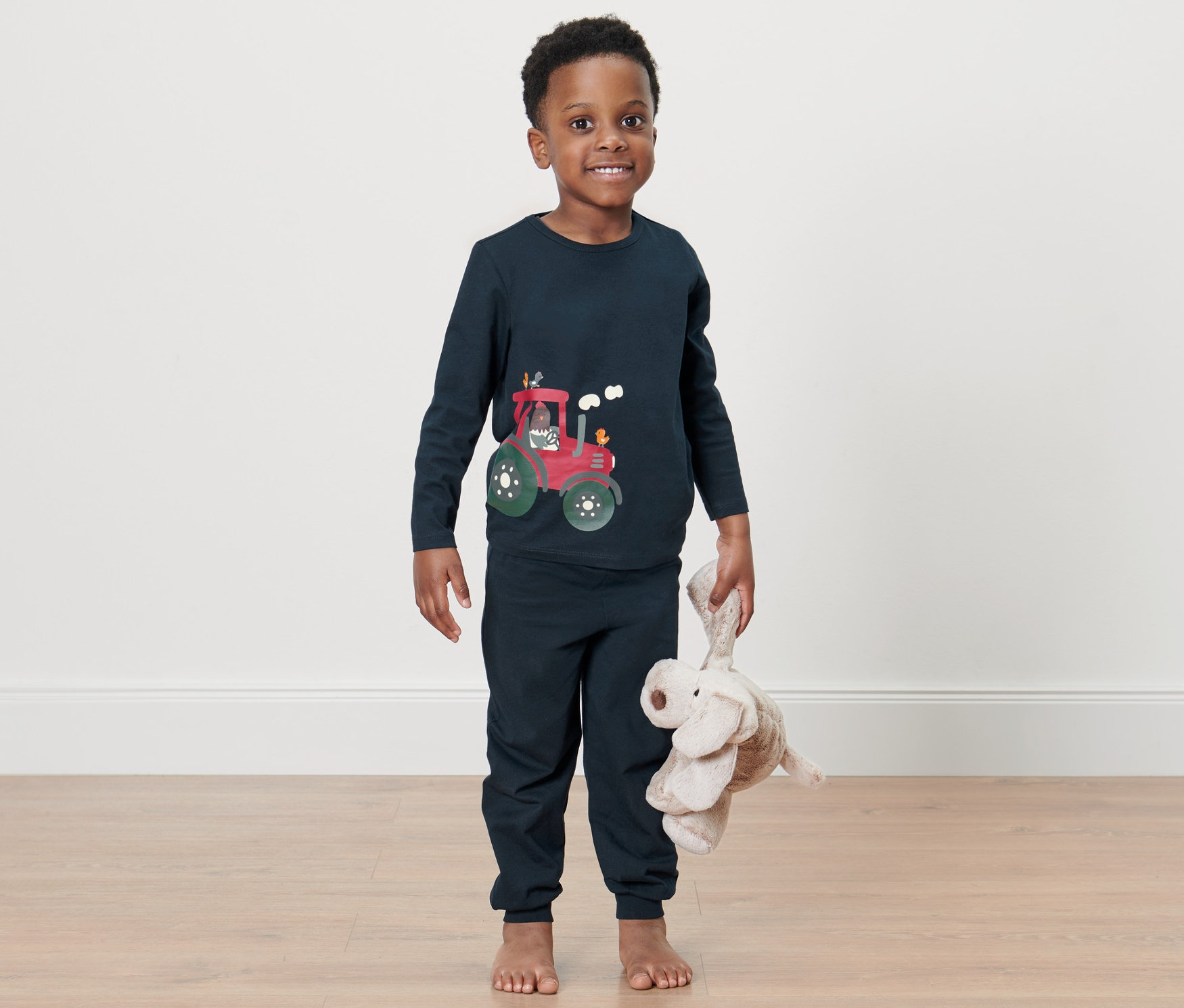 Oğlan ayakta duruyor ve peluş bir fil tutuyor. Üzerinde traktör resmi olan Küçük Çocuk Pijama Takımı var.