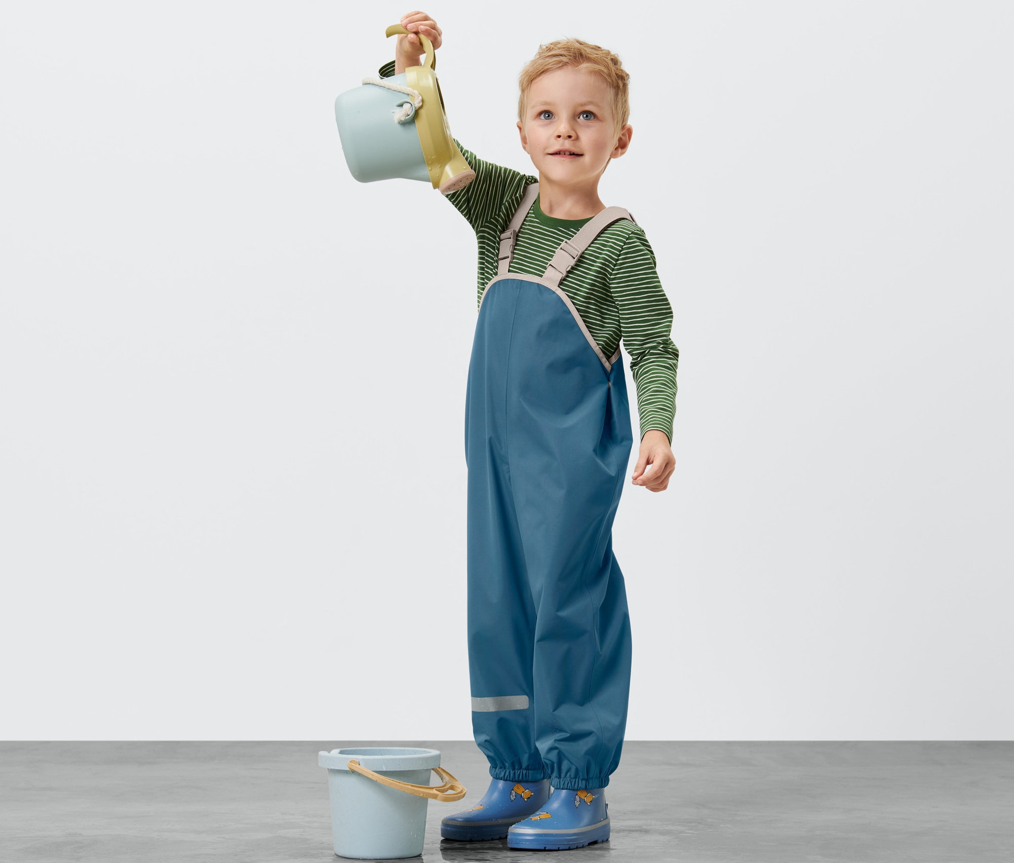 Oğlan başının üzerinde mavi bir kova tutuyor. Mavi bir Küçük Çocuk Yağmur Pantolonu giyiyor.