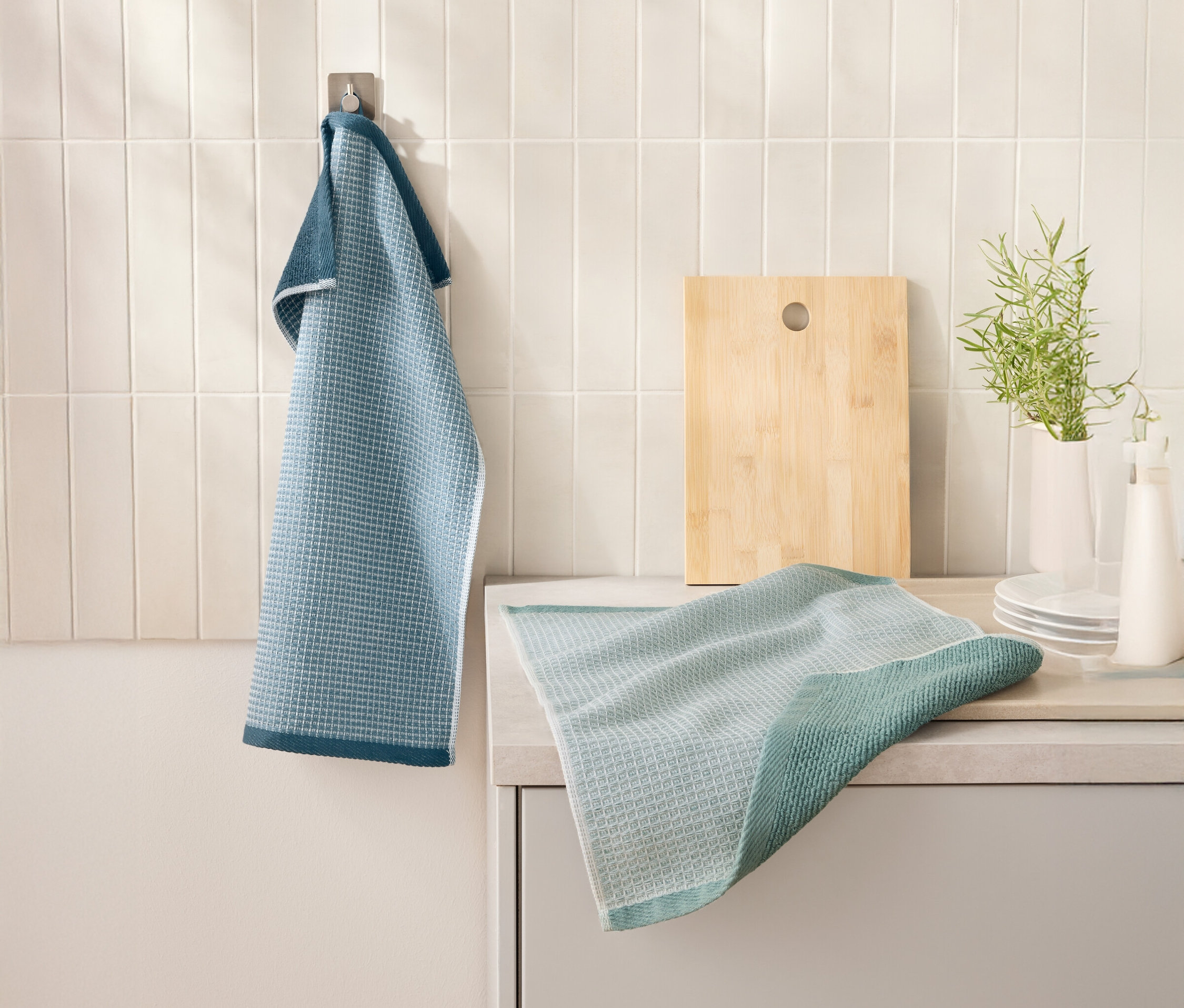 Mavi renkli 2 Waffelpiqué-Küchenhandtücher kesme tahtasının yanındaki bir kancada asılı duruyor. Başka bir mutfak havlusu mutfak tezgahında duruyor.