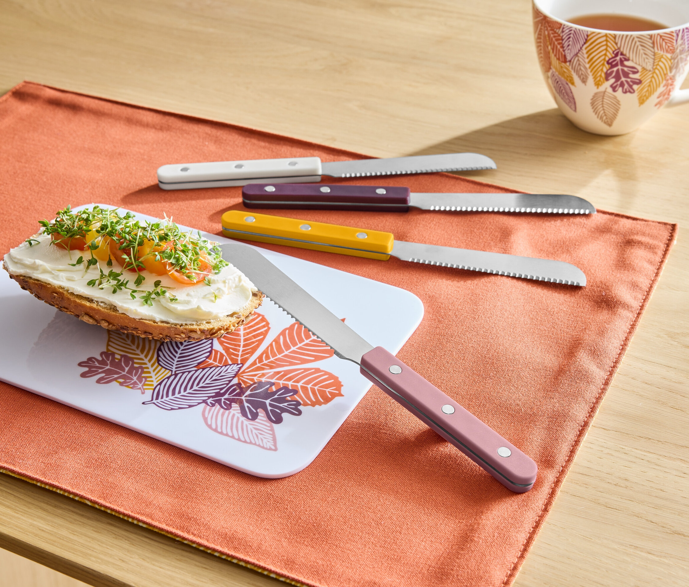 Turuncu bir Amerikan servisinde, yaprak desenli bir tahtada sürülebilir bir ekmek, yanında renkli saplı dört bıçak ve arka planda bir fincan çay duruyor.