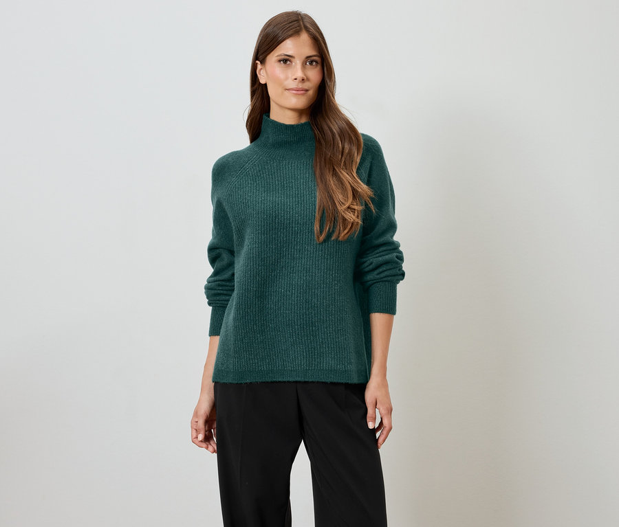 Kadın yeşil balıkçı yaka kazak ve siyah pantolonla poz veriyor.