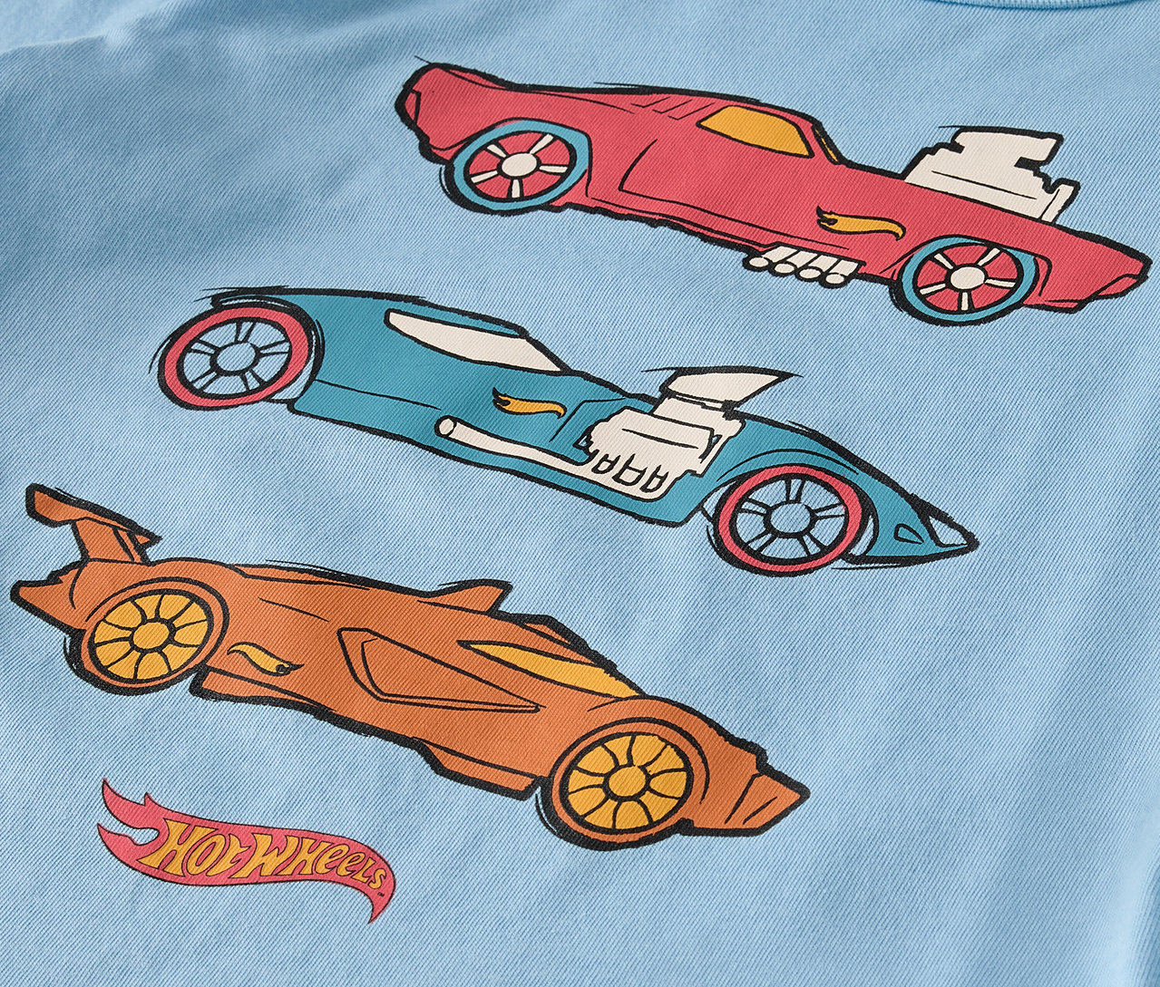 Açık mavi bir tişört üzerinde kırmızı, mavi ve turuncu renklerde üç adet çizilmiş Hot Wheels arabası bulunmaktadır.