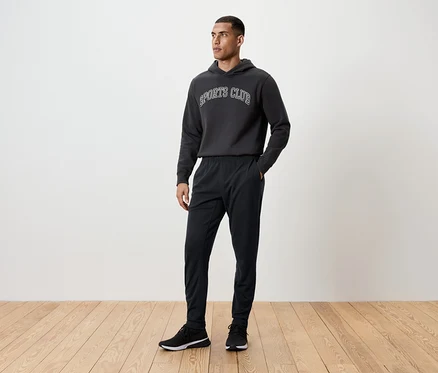 Adam ahşap bir zeminde duruyor, üzerinde siyah eşofman altı, kapüşonlu sweatshirt ve spor ayakkabısı var.