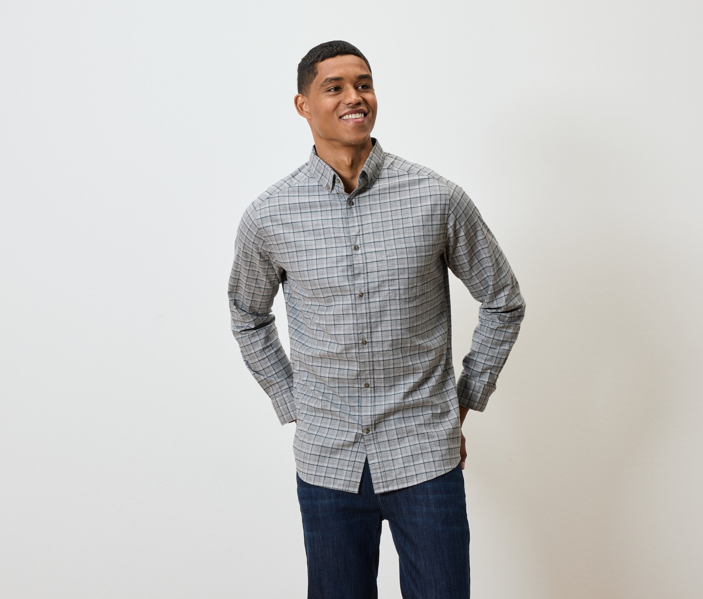 Gülümseyen bir adam gri kareli, uzun kollu bir gömlek ve koyu mavi kot pantolonla poz veriyor.