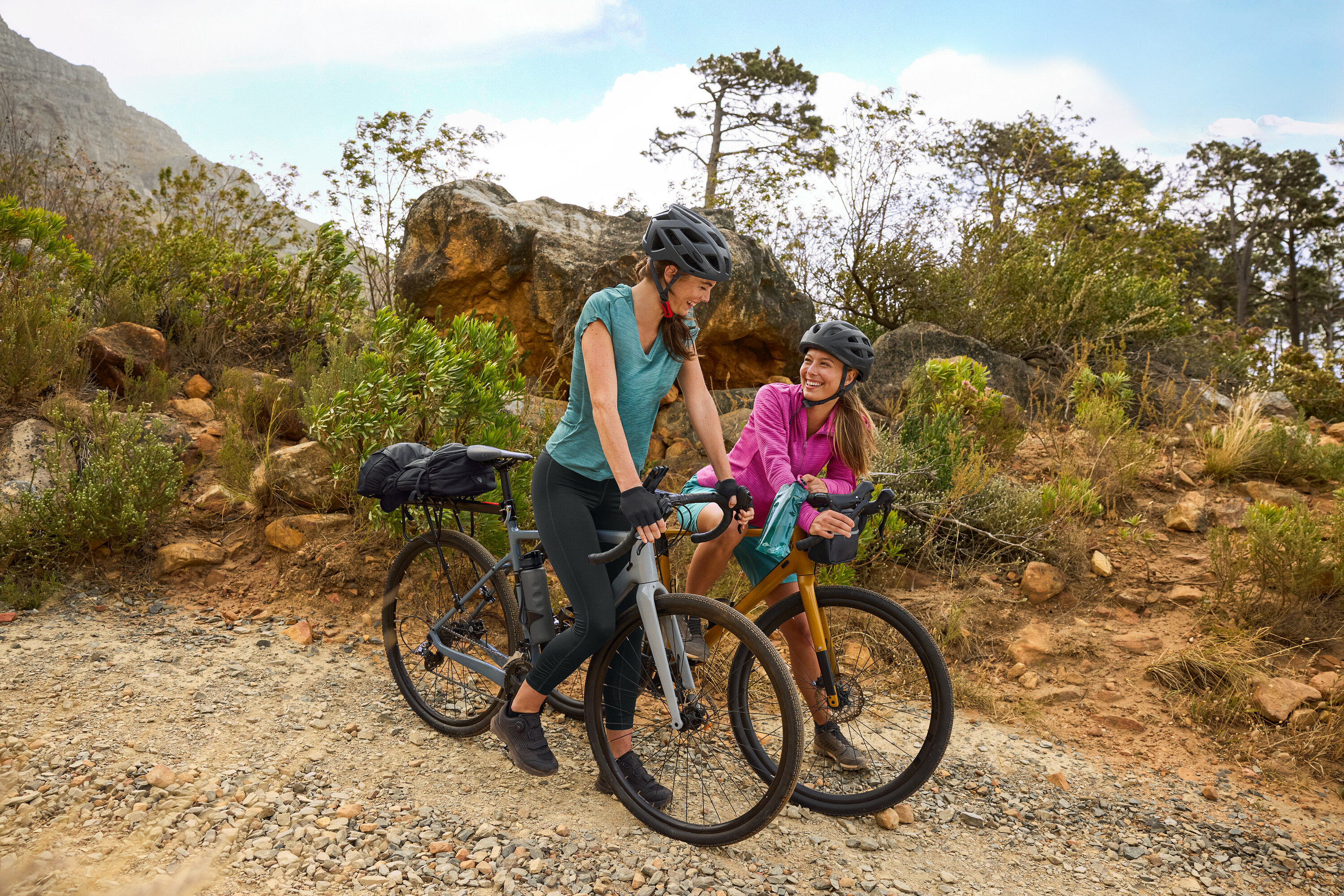 Kasklı iki kadın, engebeli arazide çakıllı bir yolda bisikletleriyle duruyor. Bir matara bisiklete takılı.