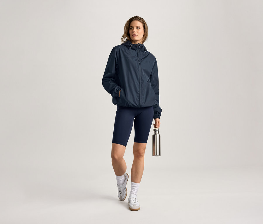 Yağmurluk, tayt ve su şişesi olan bir kadın. Çorap ve spor ayakkabı giyiyor.