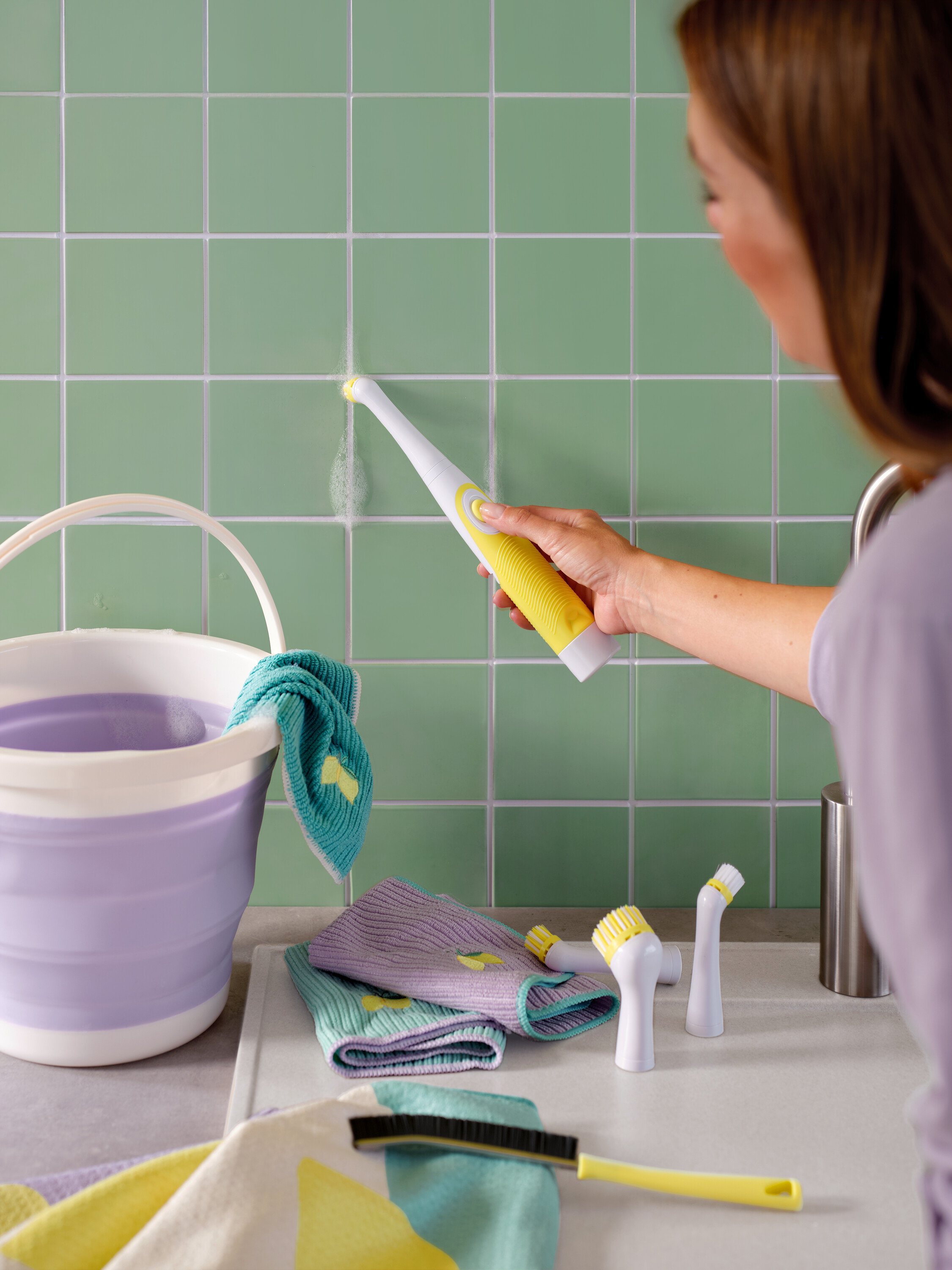 Kadın yeşil fayansları sarı-beyaz bir fırça ile temizliyor, kova ve bezler de görülebiliyor.