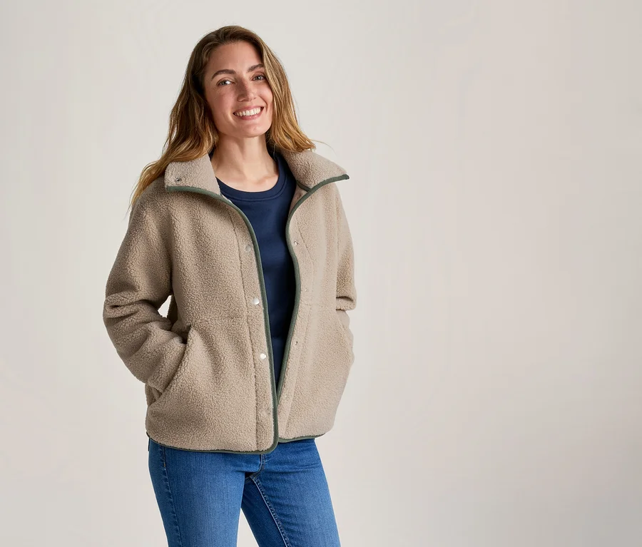 Bej rengi yeşil kenarlı polar ceket, lacivert tişört ve kot pantolon giyen, elleri ceket ceplerinde gülümseyen bir kadın.