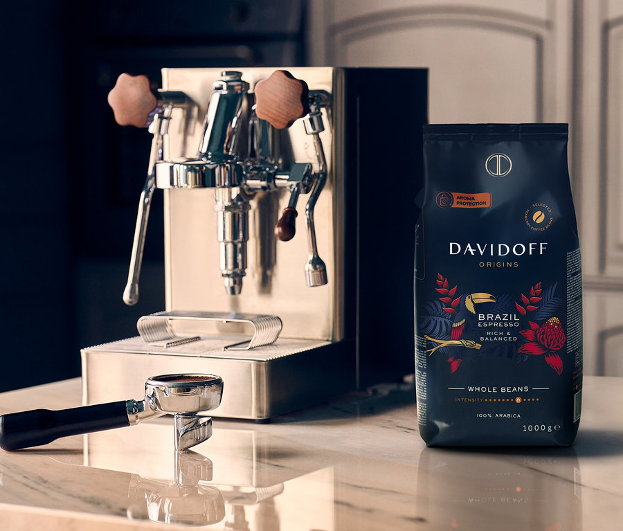 Mermer bir yüzeyde, bir kahve makinesi DAVIDOFF CAFÉ ORIGINS Brazil - 1x 1000 g Bütün Çekirdek kahve paketinin yanında duruyor.