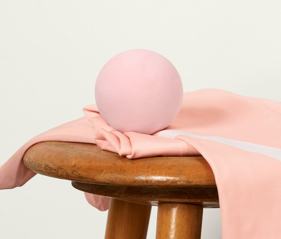 Pembe bir fasya masajı topu, ahşap bir tabure üzerinde duran beyaz şeritli pembe bir taytın üzerinde duruyor. Arka planda pembe bir yoga matı var.