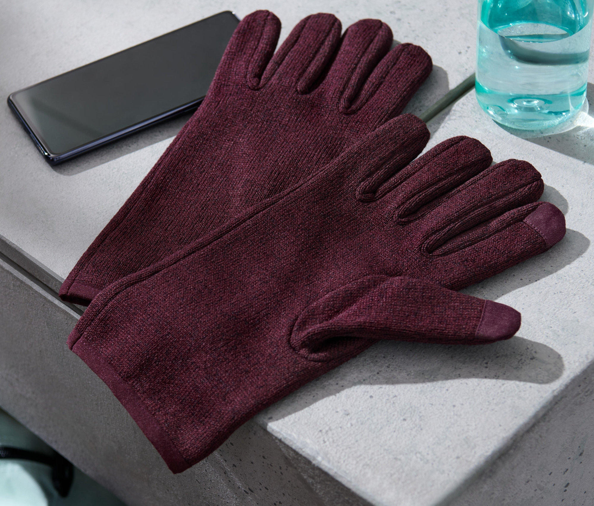 Gri bir yüzeyde örgü polar eldivenler, bir cep telefonu ve bir su şişesi duruyor.