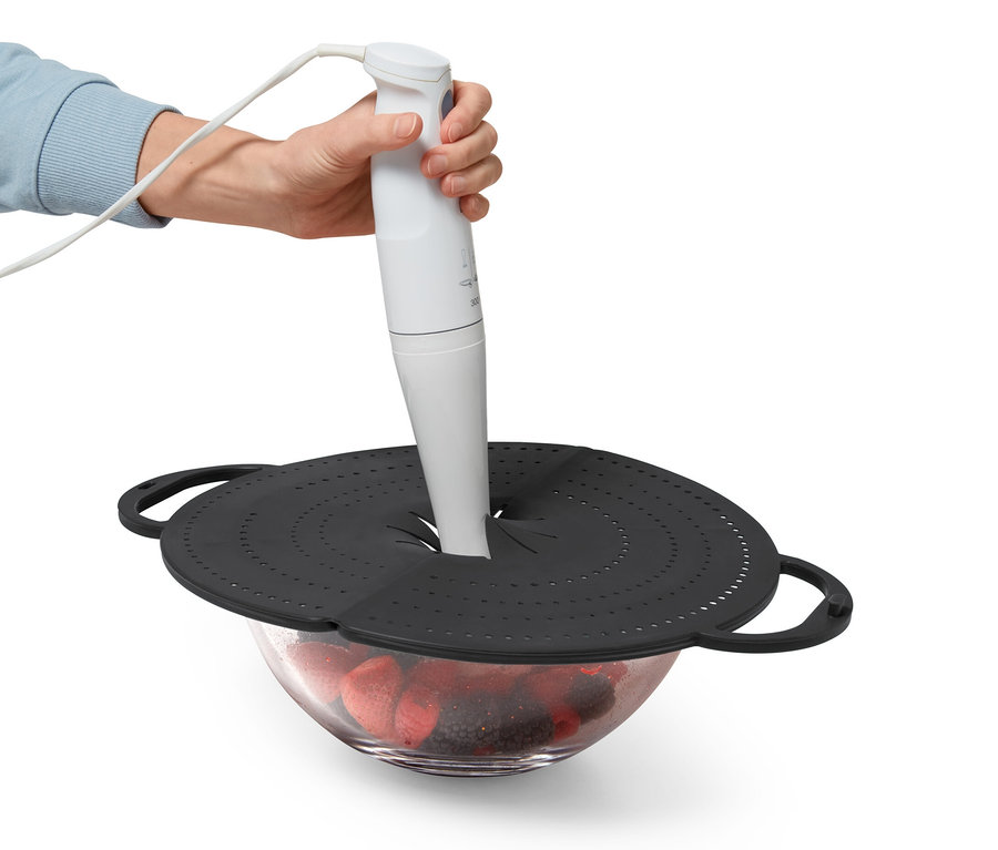 Bir el, meyvelerle dolu bir cam kâsenin üzerindeki siyah Yağ Sıçratmazdan bir blender tutuyor.