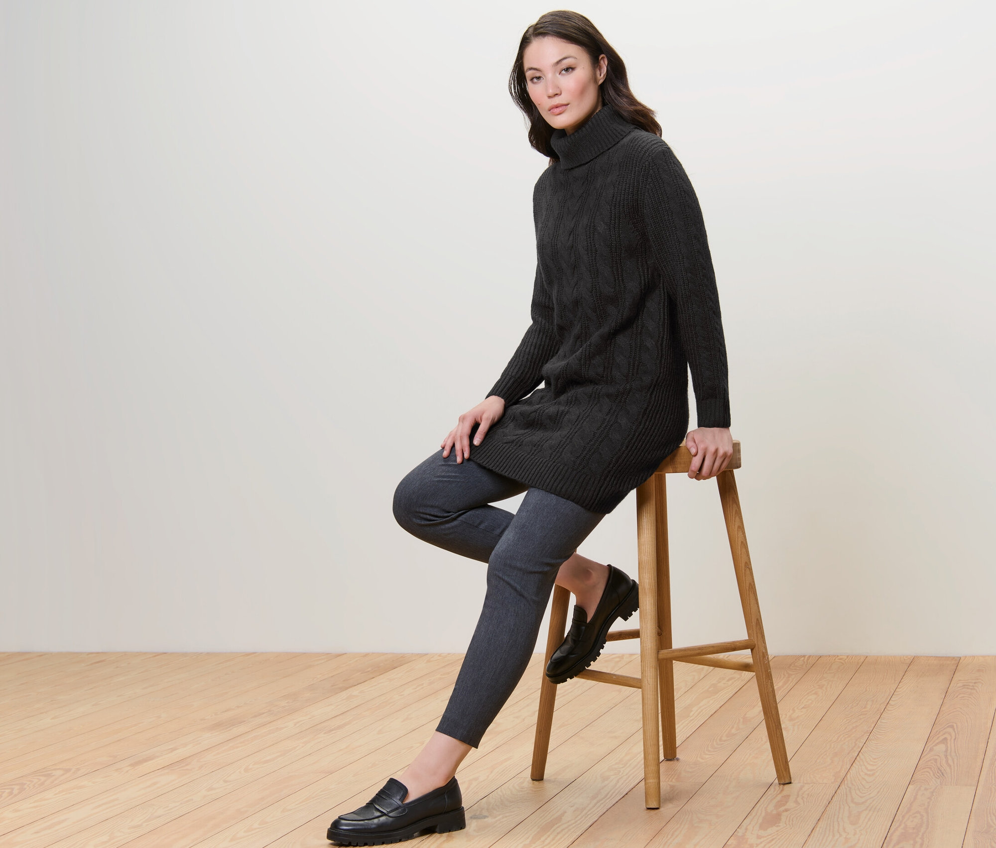 Kadın bir taburede oturuyor ve Saç Örgüsü Motifli Uzun Siyah Örgü Kazak giyiyor.