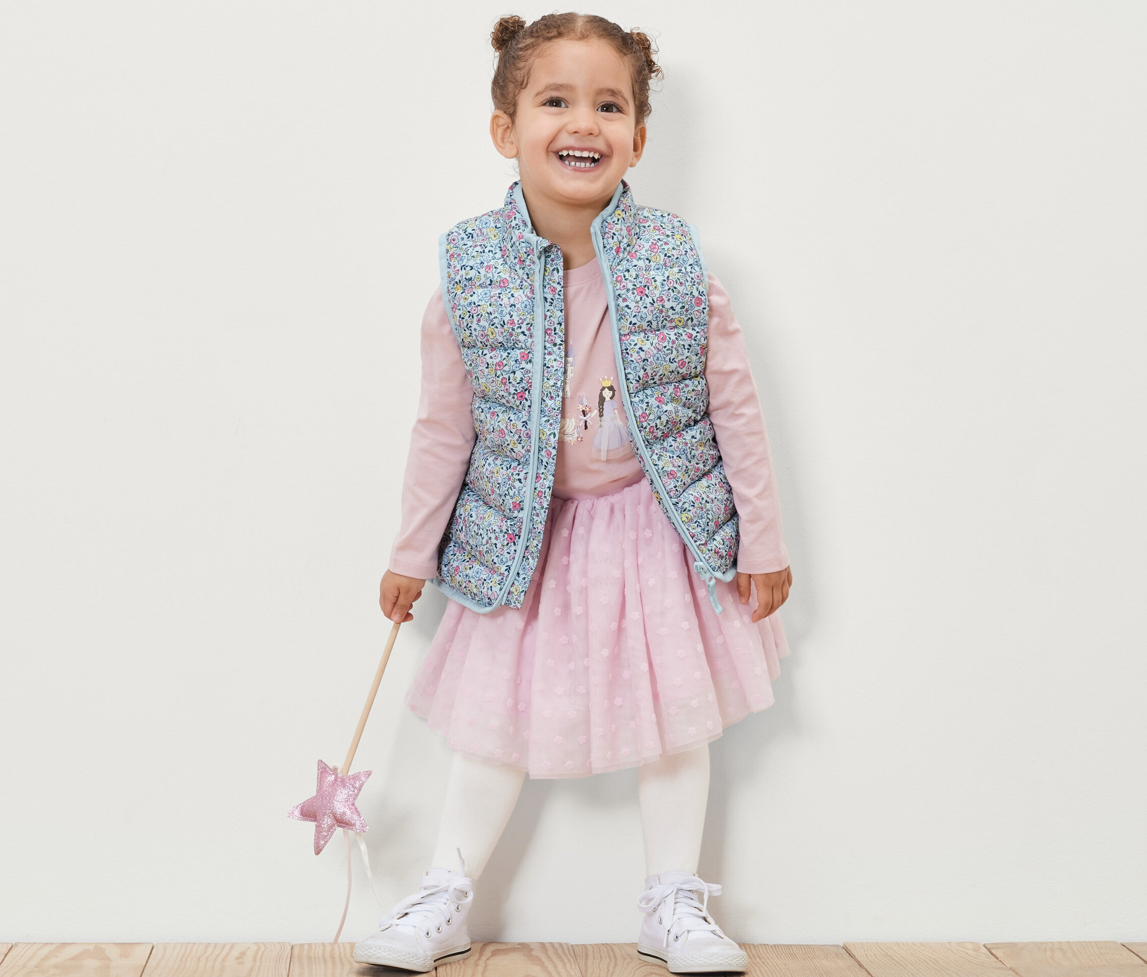 Gülümseyen küçük kız, çiçekli çocuk kapitone yelek, pembe uzun kollu tişört, pembe etek ve beyaz spor ayakkabı giyiyor ve bir sihirli değnek tutuyor.
