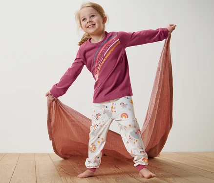 Gülümseyen kız, ahşap zeminde duruyor ve pembe çocuk pijama takımını tutuyor.