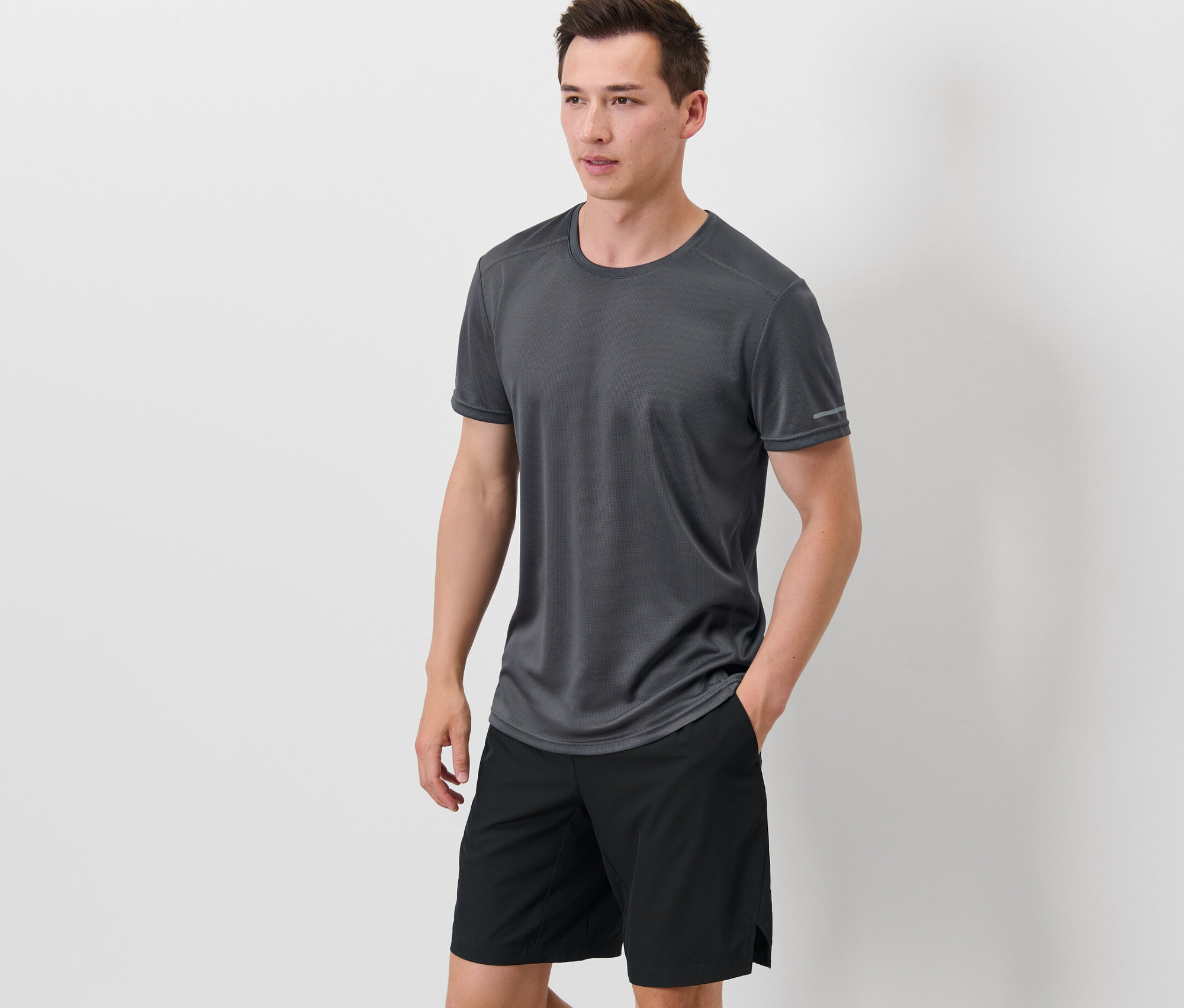 Adam siyah şortunun ceplerinde elleriyle duruyor, üzerinde gri fonksiyonel tişört var.