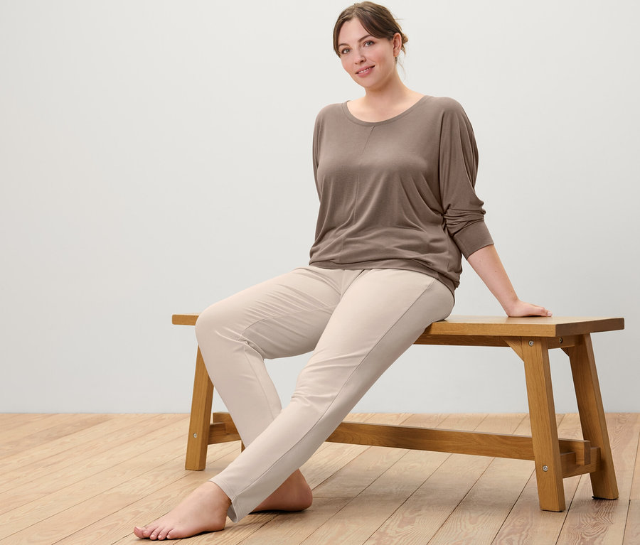 Bir bankta oturan kadın, boz renkli spor ve yoga tişörtü ve bej renkli eşofman altı giyiyor.