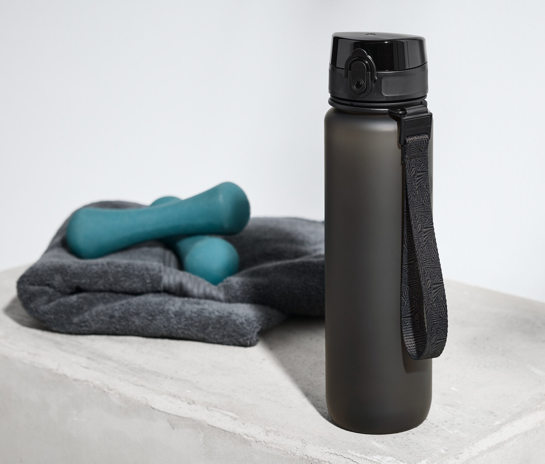 Siyah matara, gri bir yüzeyde havlu ve dambılların yanında duruyor.