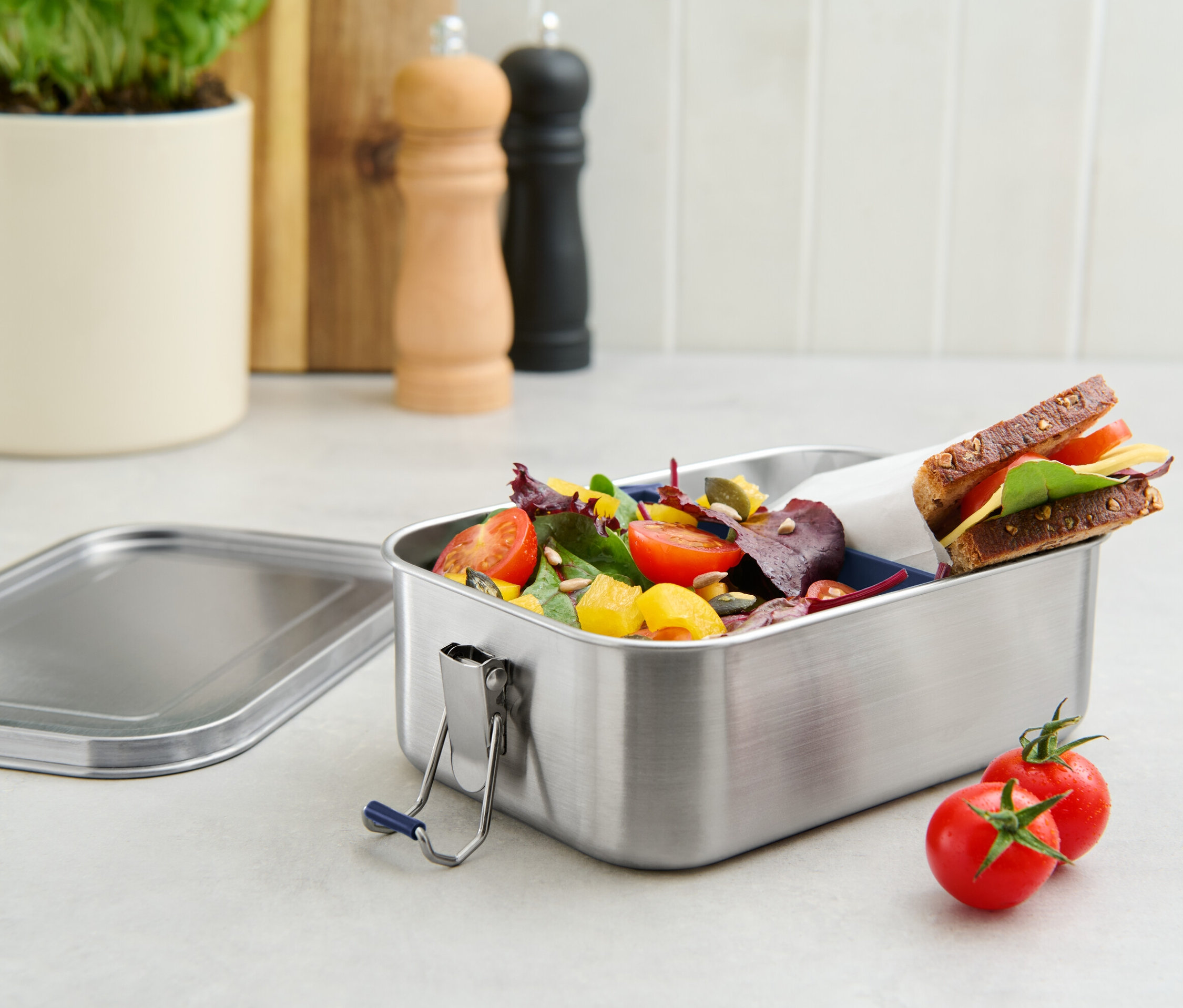 Salata ve sandviç ile dolu paslanmaz çelik taşınabilir yemek kutusu, yanında iki domates var.