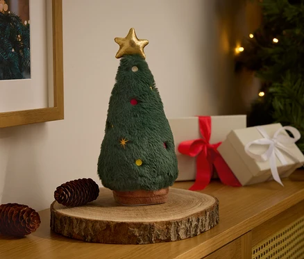 Altın yıldızlı dekoratif pelüş Noel ağacı, iki çam kozalağı ve hediye kutularının yanında ahşap bir taban üzerinde duruyor.