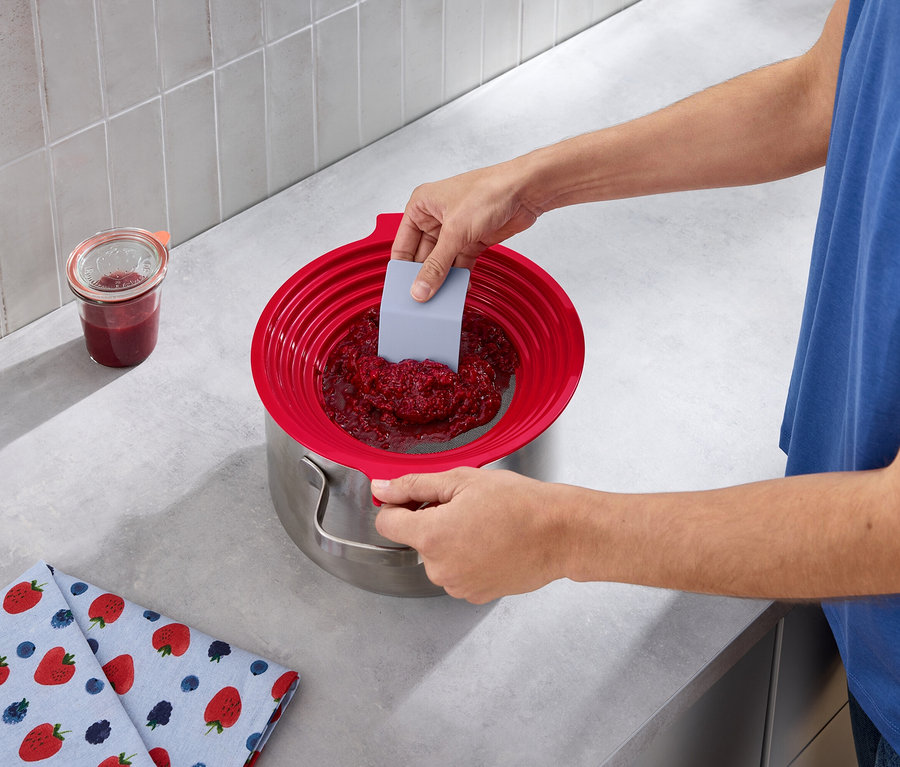 Adam, gri bir spatula yardımıyla bir tencereye 2'si 1 arada un ve püre eleğinden ahududu reçeli geçiriyor. 