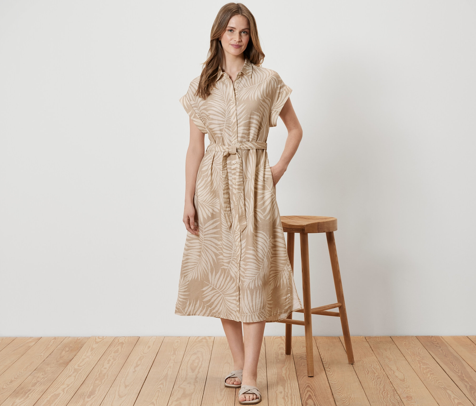 Kadın ahşap taburenin yanında duruyor ve baskılı bir keten elbise giyiyor.