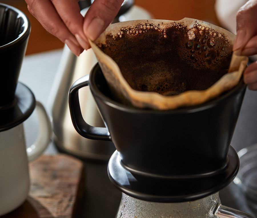 Kadın, siyah Kaffee Handfilter Gr. 2'nin içine kağıt filtre yerleştiriyor. Altında Kaffeekanne, Glas bulunuyor.