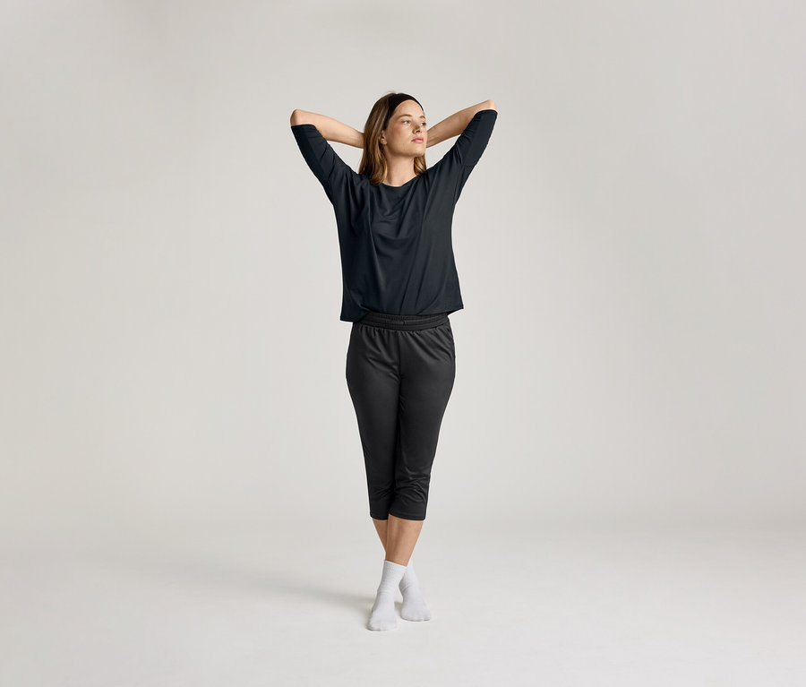 Elleri başının arkasında kenetli olan kadın, siyah renkli 3/4 spor tişörtü giyiyor.