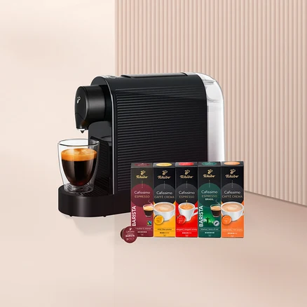 Siyah beyaz kapsül kahve makinesi, altında cam bir fincan kahve ve yanında beş kutu Tchibo Cafissimo kahve kapsülü ile bir adet Barista kapsülü.