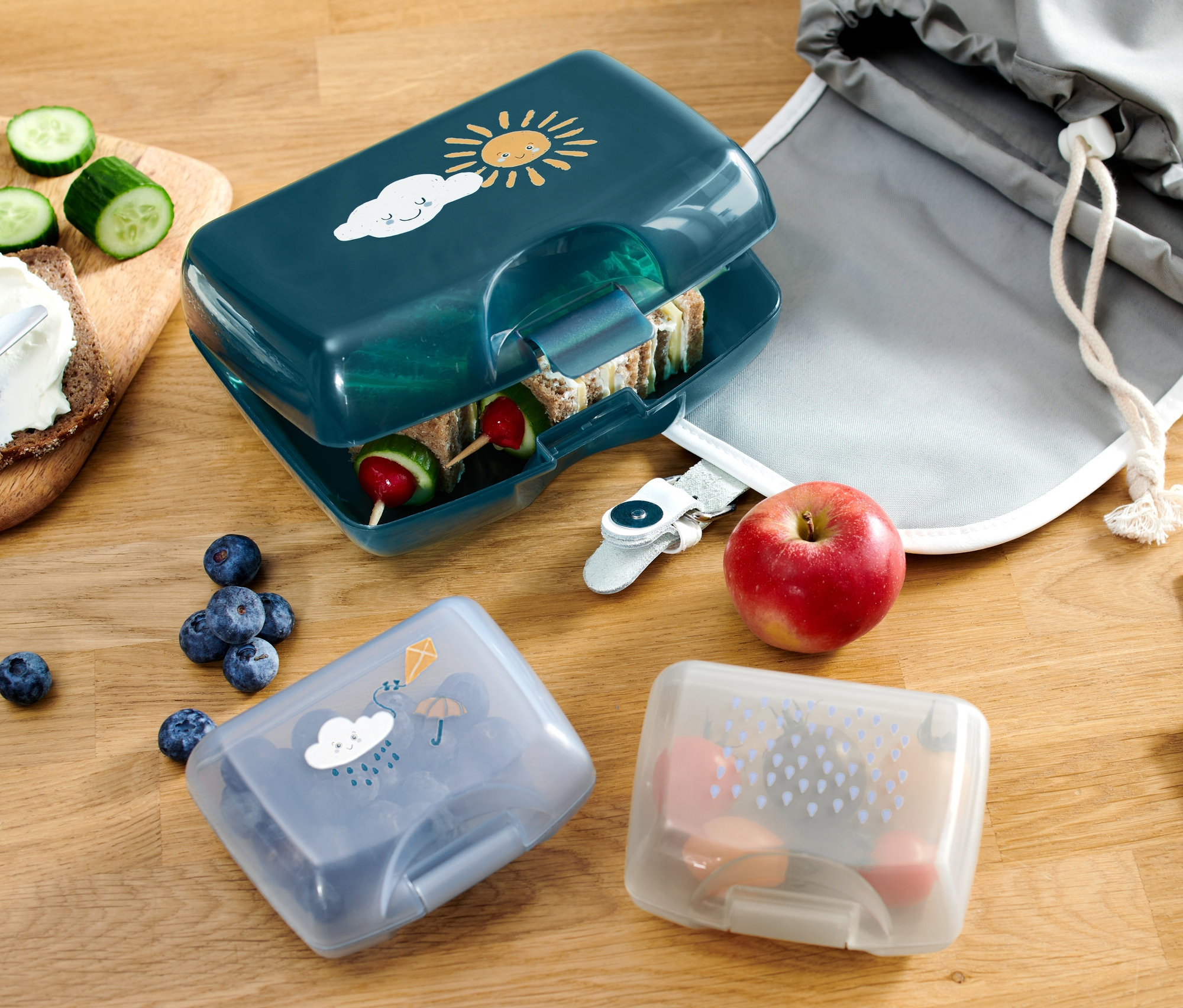 Masa üzerinde açık duran, güneş ve bulut desenli, mavimsi yeşil renkte 3 Adet Beslenme Kutusu, Yağmur, yağmur desenli 2 adet küçük, kapalı 3 Adet Beslenme Kutusu, Yağmur, bir elma, yaban mersinleri, sürülebilir ürünlü ekmek, gri kumaş çanta ve boya kalemleriyle birlikte bir defter bulunmaktadır.