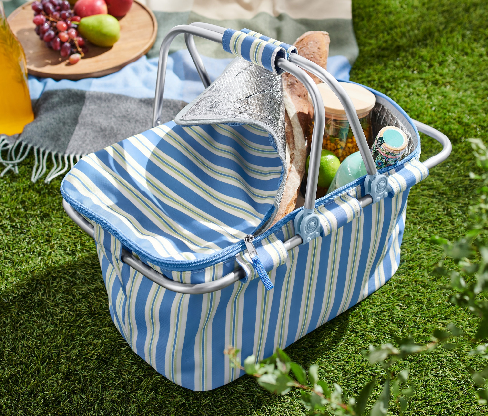 Çimenlerin üzerinde açık bir soğutucu sepet, yanında elma, üzüm ve içecek bulunan bir örtü.