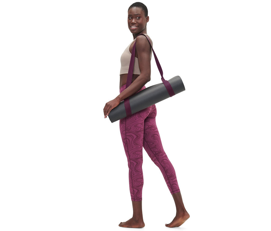 Kadın omzunda gri bir yoga matı tutan 2'si 1 Arada Yoga Kemeri ile yürüyor.