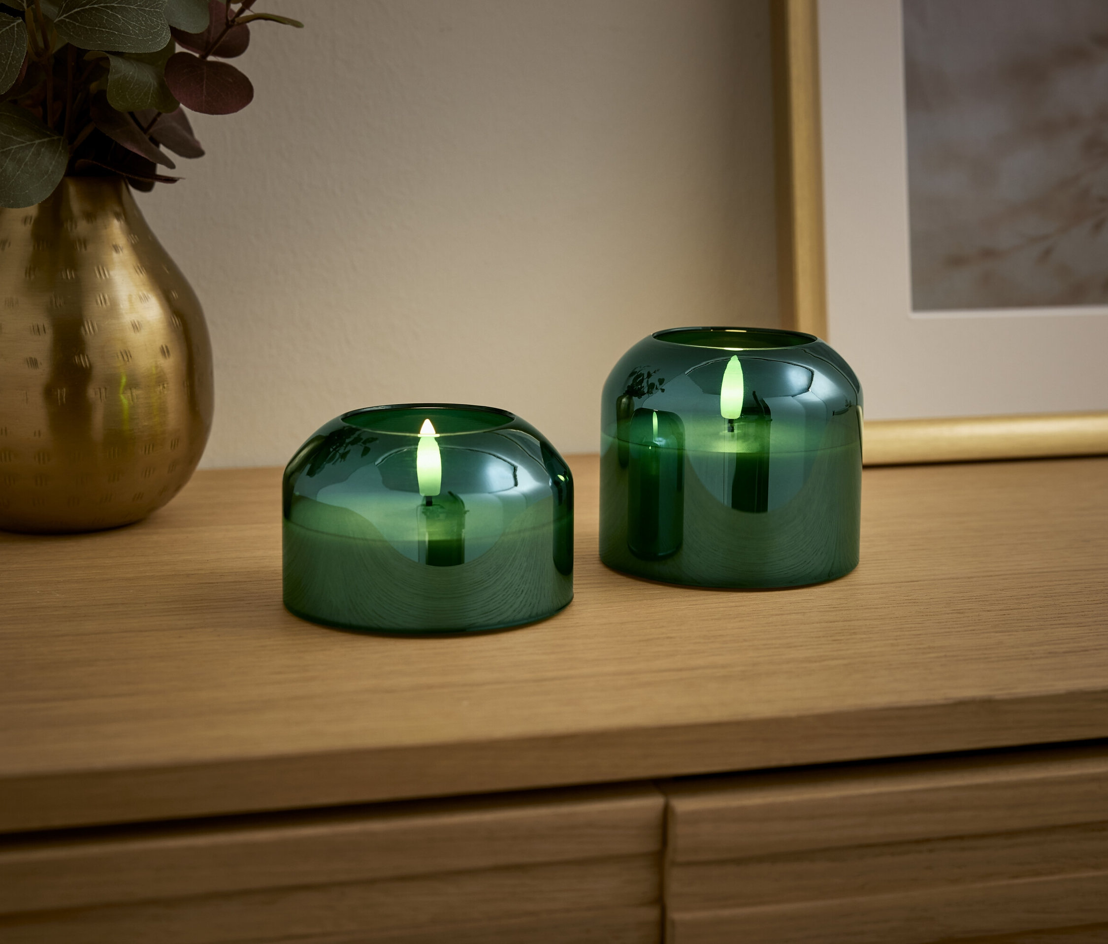 Ahşap bir masa üzerinde iki adet yeşil mum ve altın bir vazo bulunmaktadır.