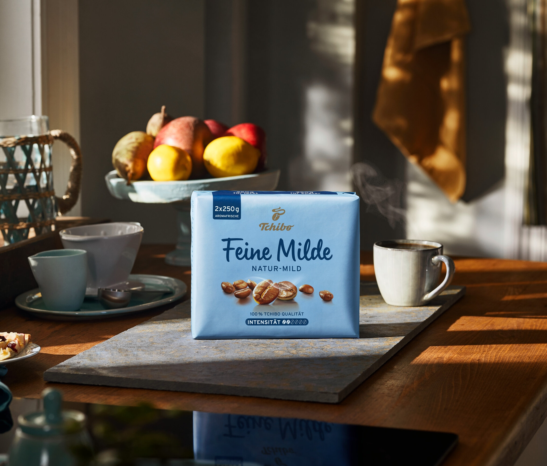 Bir paket Feine Milde Öğütülmüş Filtre Kahve, ahşap bir masanın üzerinde bir fincan sıcak kahvenin yanında duruyor.